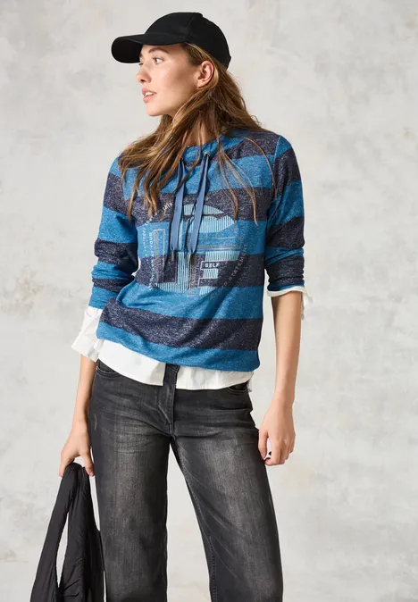 Frau mit blau gestreiftem Pullover, schwarzer Kappe und Jeans schaut zur Seite.